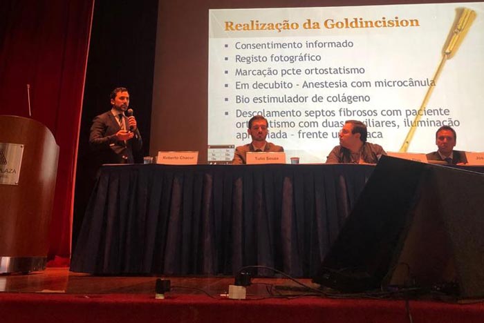 médico em pé no palco do congresso mundial de medicina estética ao lado de mesa com outros médicos e projeção ao fundo de como é realizada a técnica goldincision de tratamento para celulite