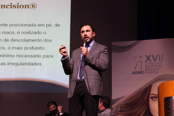 homem branco barba por fazer de terno com microfone na mão em cima do palco com projetor atrás